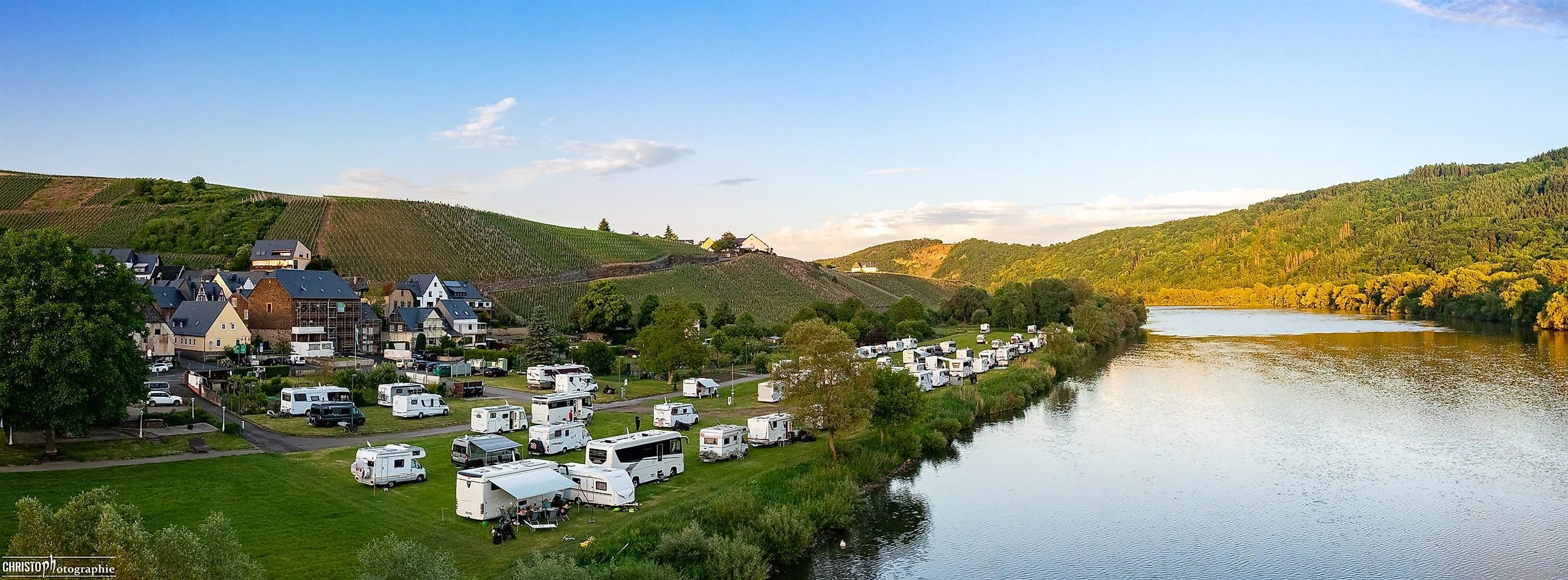 Eine malerische Landschaft entlang eines Flusses mit Campingplätzen und grünen Hügeln. Der Himmel ist klar und die Sonne scheint.